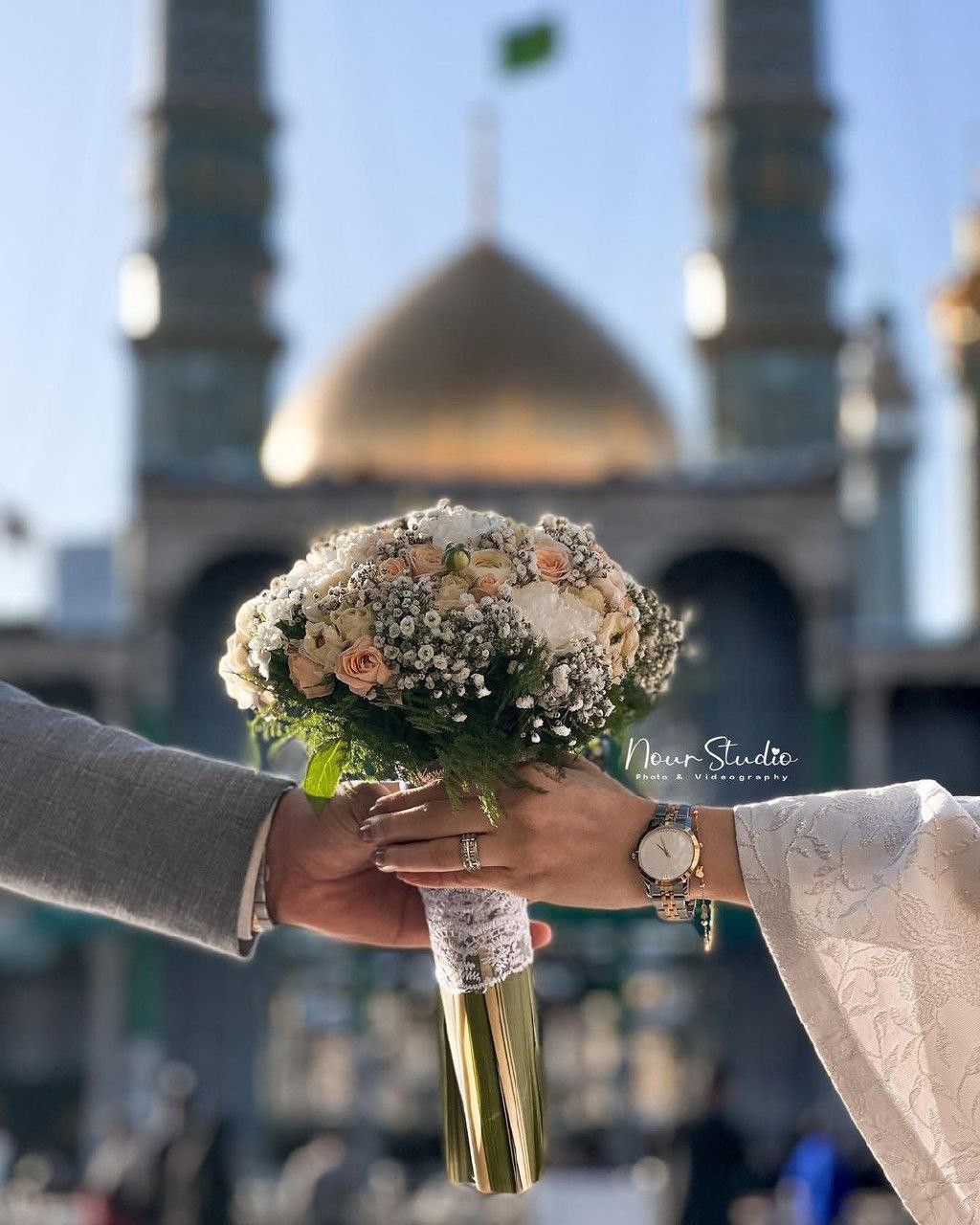 دست عروس و داماد دسته گل رز کرم و هلویی را مقابل گنبد طلایی و پرچم سبز حرم نگه داشته‌اند