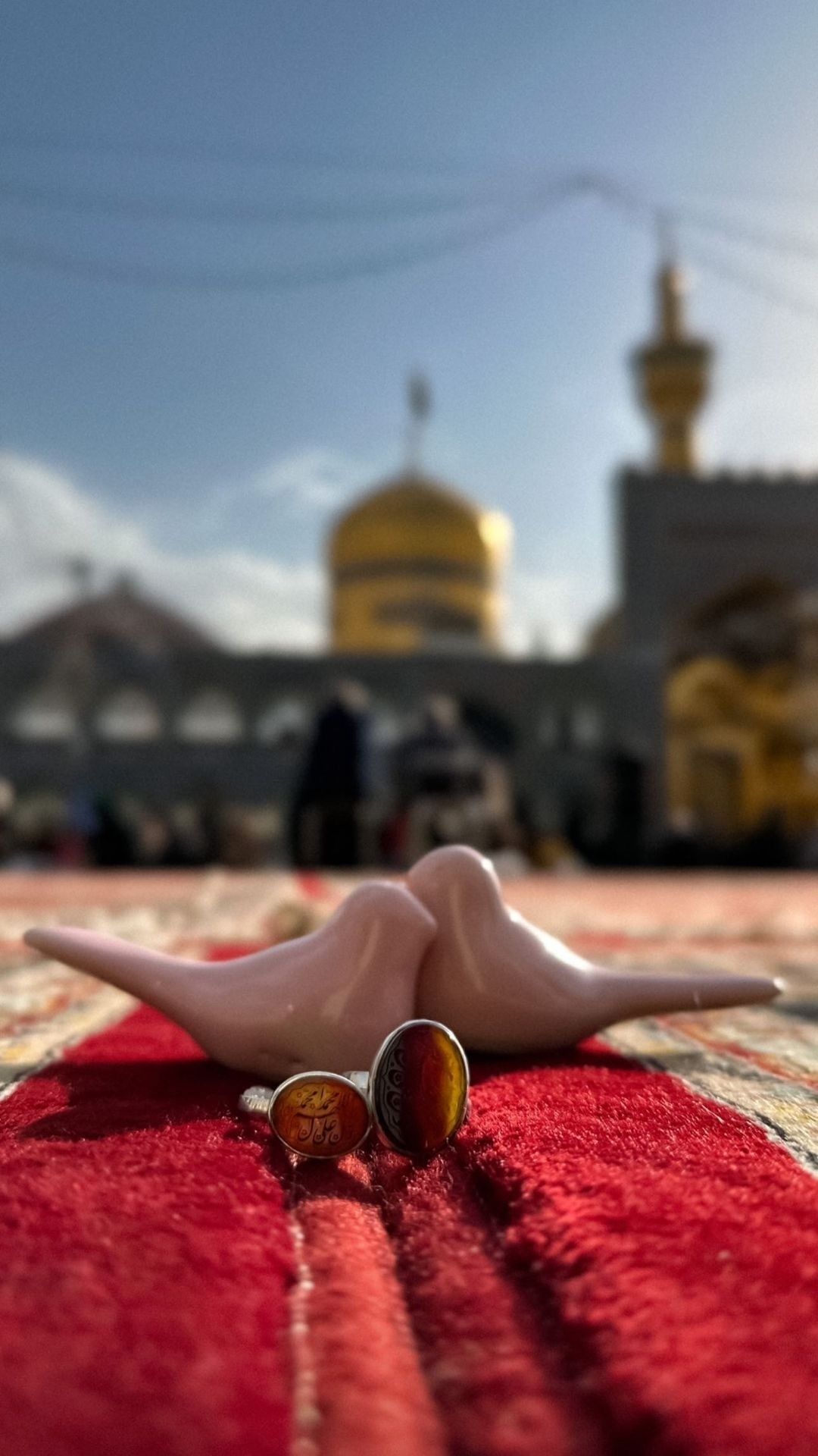 دو پرنده سفالی صورتی و دو انگشتر عقیق روی فرش سرخ با گنبد طلایی حرم در پس‌زمینه