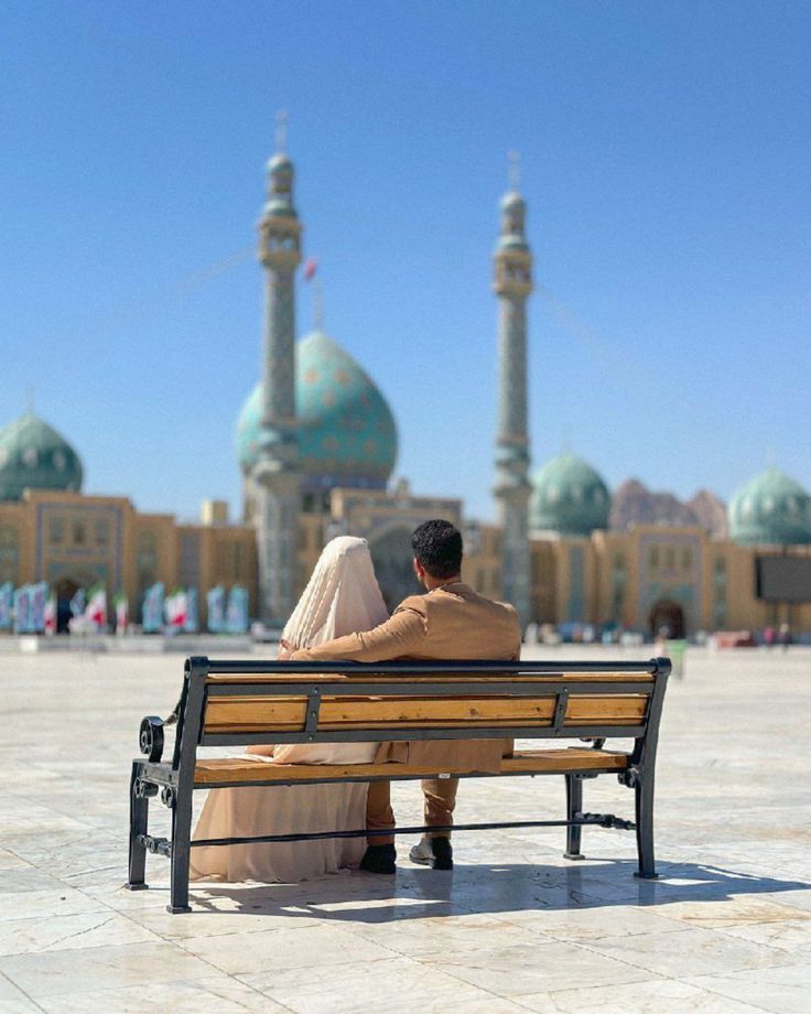 عروس با چادر کرم و داماد با کت شتری روی نیمکت چوبی رو به مسجد با گنبد فیروزه‌ای نشسته‌اند