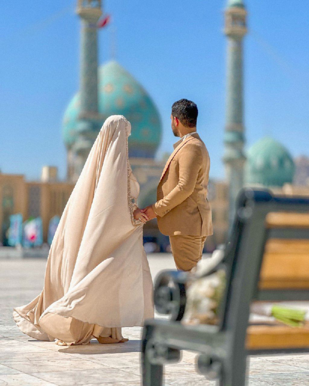 عروس با چادر کرم و دنباله بلند و داماد با کت شتری دست در دست در صحن مسجد با گنبد فیروزه‌ای