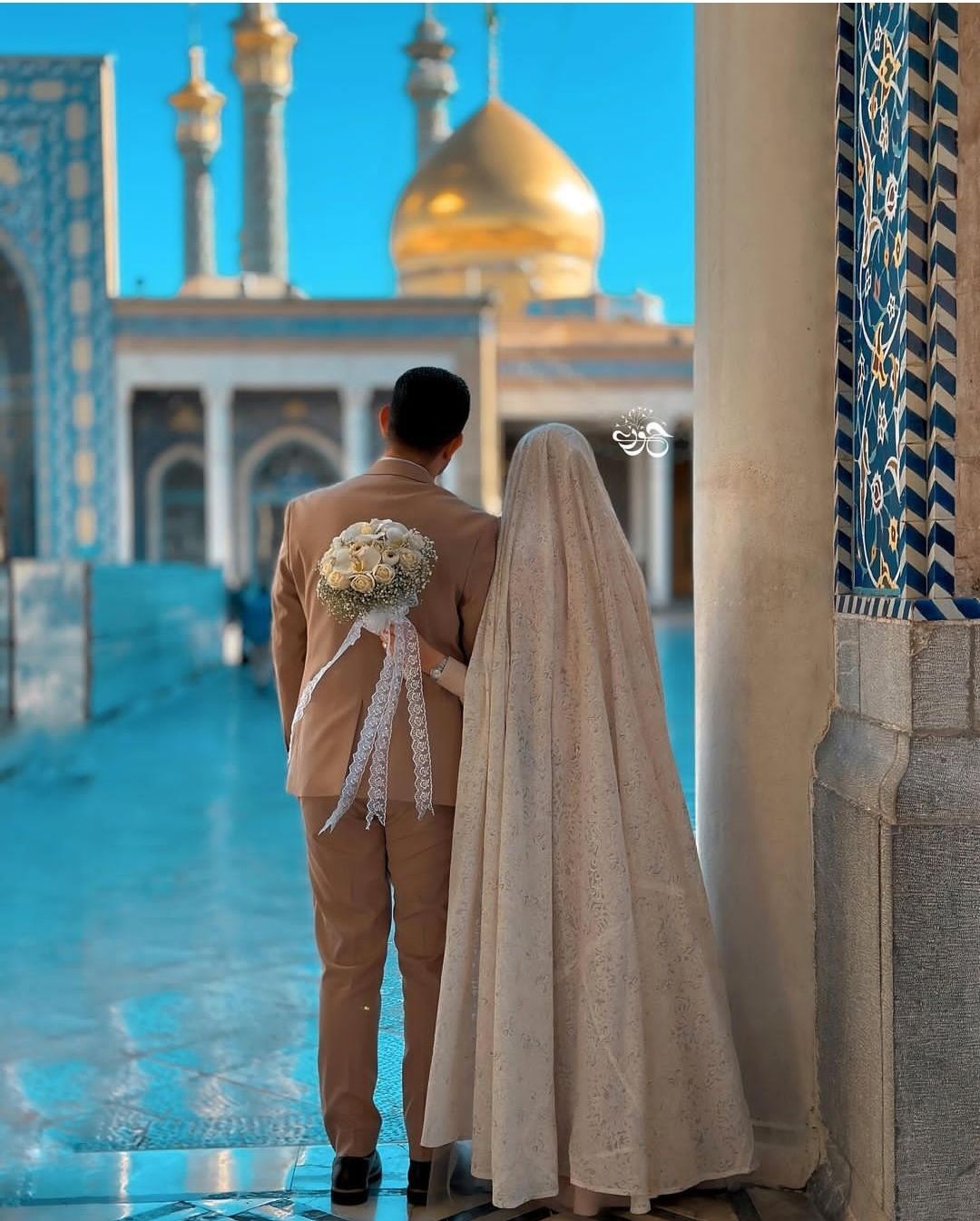 داماد با کت شتری و دسته گل رز سفید و عروس با چادر کرم مقابل گنبد طلایی و صحن فیروزه‌ای