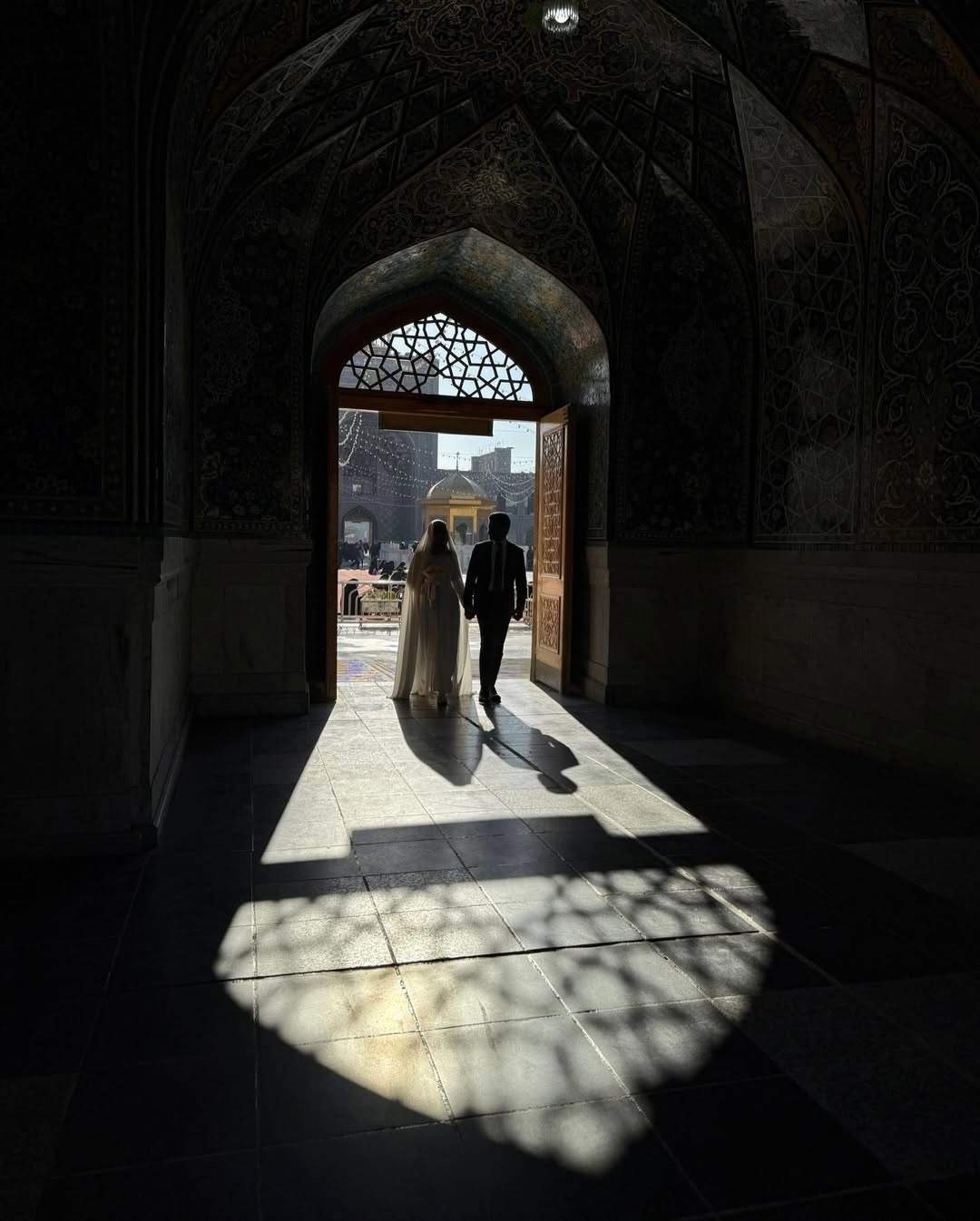 زوجی دست در دست از در چوبی مسجد بیرون می‌روند و نور مشبک روی کف سنگی افتاده است