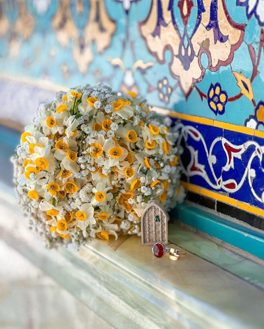 دسته گل نرگس و ژیپسوفیلا روی لبه کاشی فیروزه‌ای کنار ماکت ضریح و انگشتر عقیق سرخ
